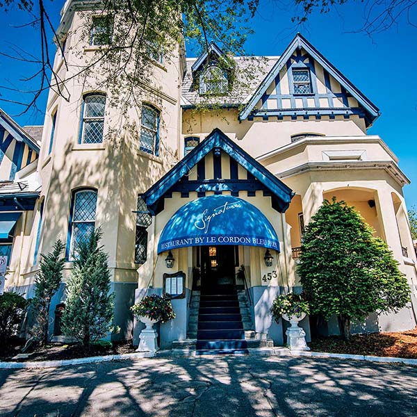 The cream-coloured heritage building exterior of Le Cordon Bleu Ottawa, with a blue awning reading "Restaurant by Le Cordon Bleu" over the entrance.