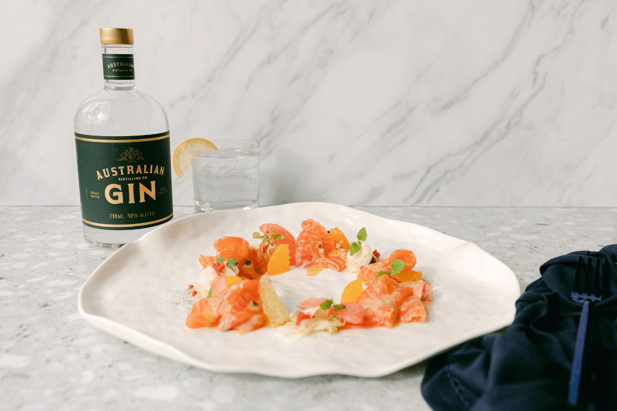 salmon ceviche on white plate and, left, bottle of Australian Distilling Co Gin on a marble table with navy napkin and fork to the right.