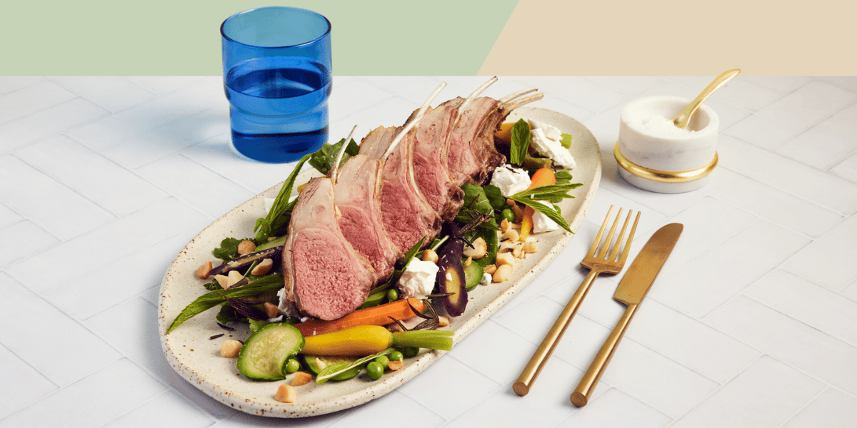 smoked lamb rack on a stone oval plate, surrounded by gold cutlery, salt bowl and blue glass of water on a mint green and yellow background.